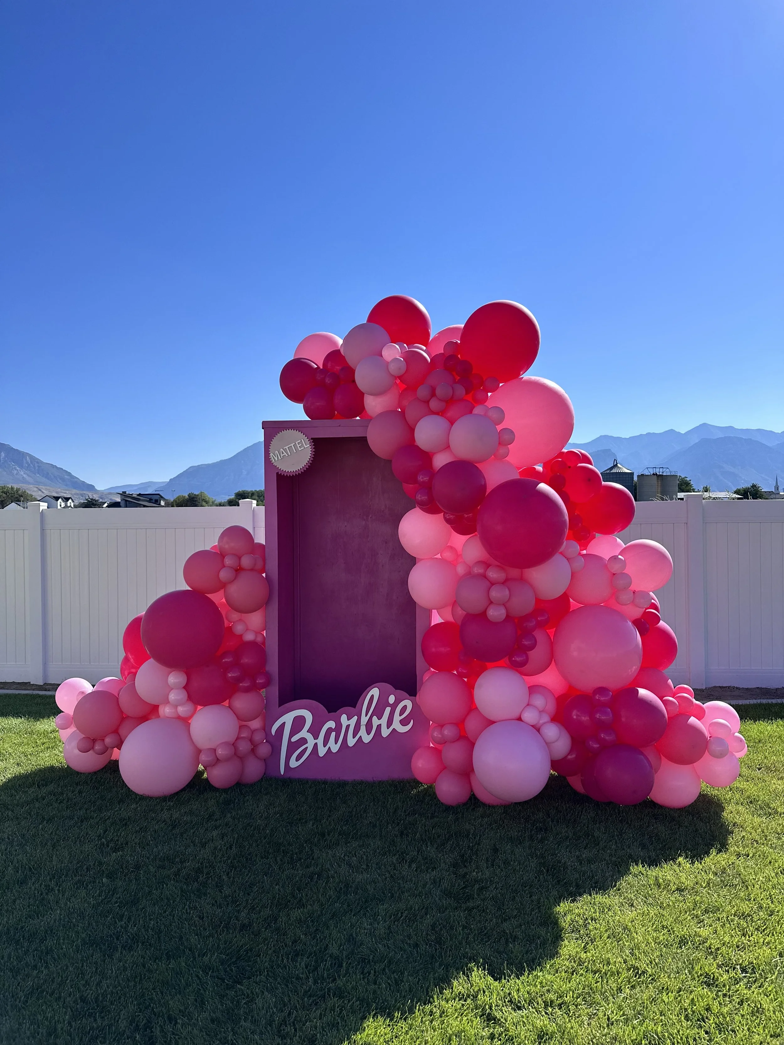 Transport into the world of Barbie: A life-size Barbie box adorned with vibrant pink balloons stands on lush green grass in a backyard. A fun celebration of the Barbie movie, with a photo backdrop inviting guests to step inside the box and immerse th