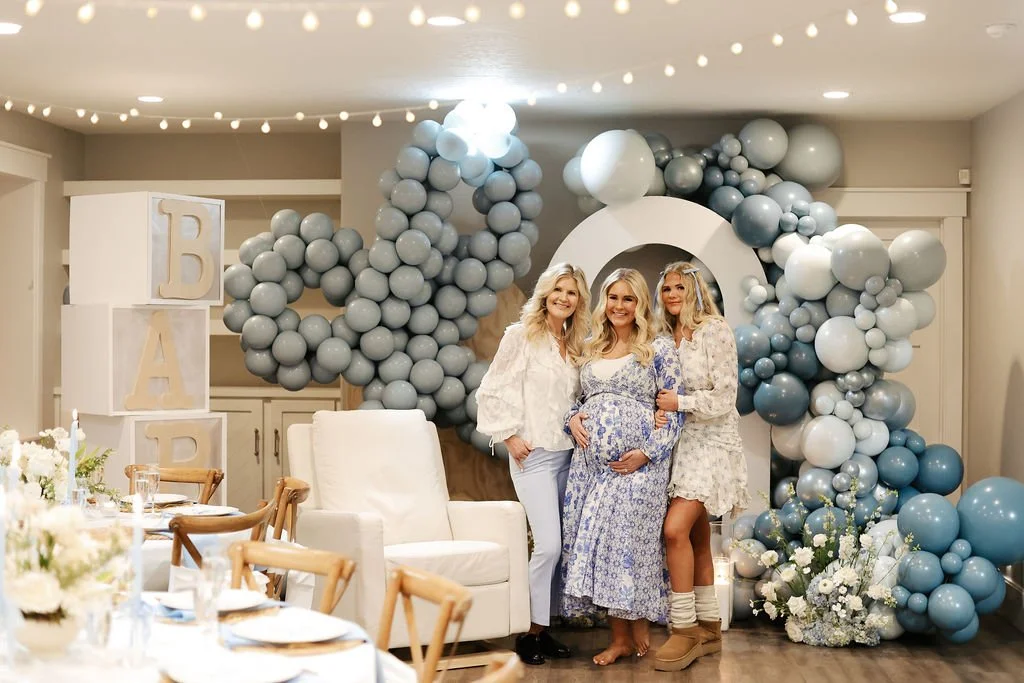 Charming baby shower for a baby boy featuring elegant white arches adorned with blue and white balloons. A giant blue bow and oversized baby blocks spell 'Baby'. Three joyful women, including the mom-to-be, stand proudly before the lovely display.