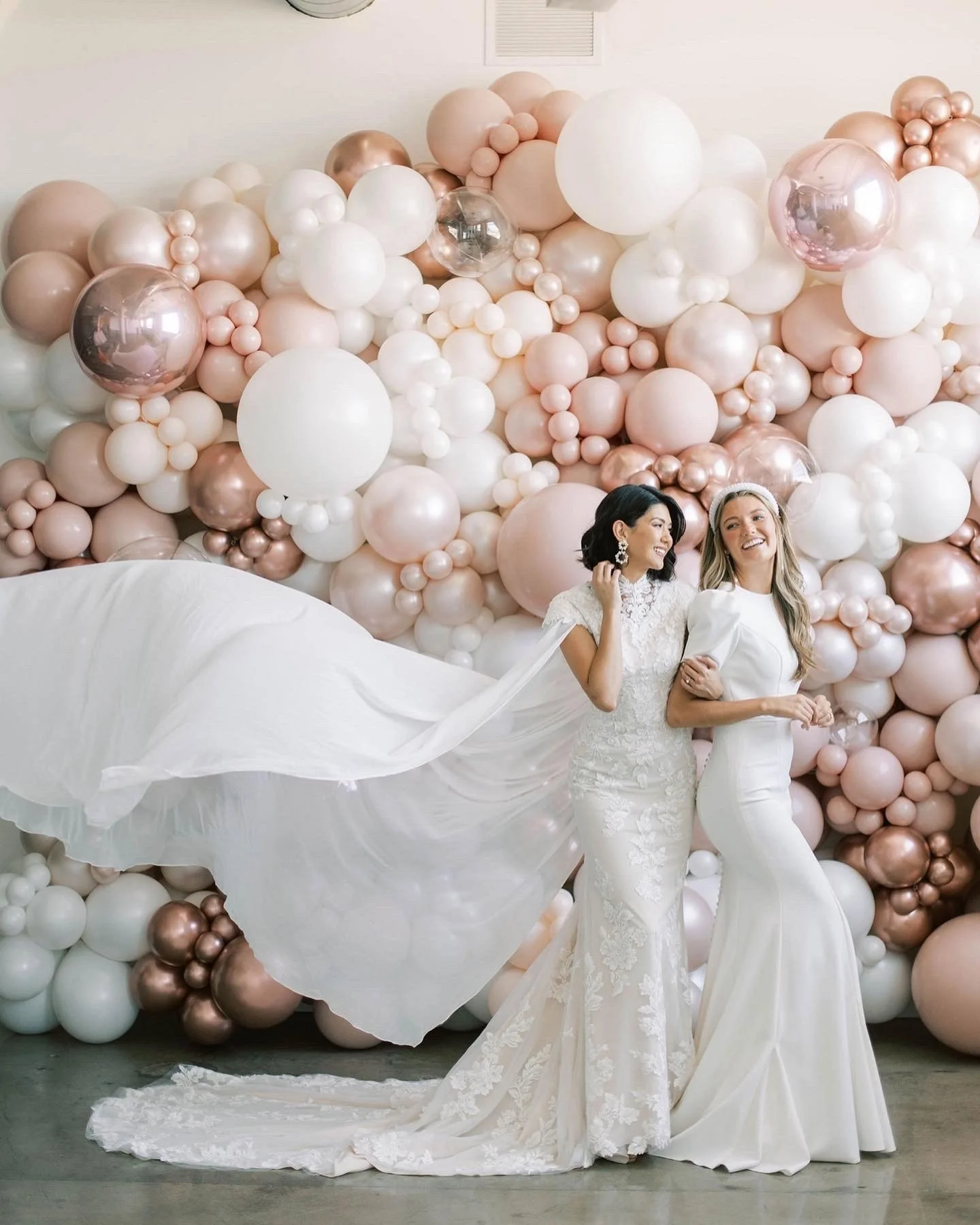 Utah Valley Bride Magazine styled shoot: Two smiling brides, one with dark hair in a cape gown, the other blonde with a pearl-beaded headband, stand before a blush, rosegold, white, and clear balloon wall, radiating joy.