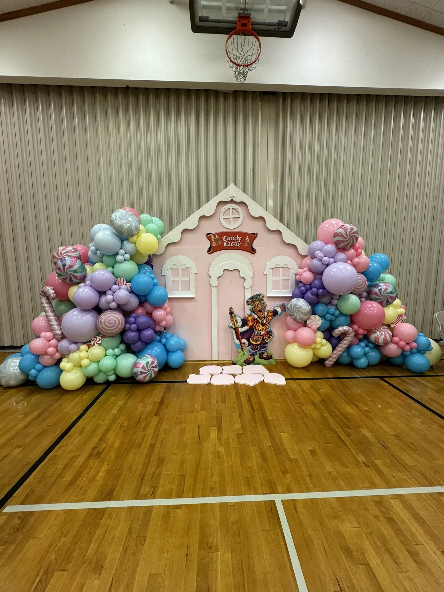 Whimsical Candyland birthday party: A charming pink "Candy Castle" backdrop adorned with faux frosting sets the scene. Pastel balloons frame the castle, accompanied by delightful candy-shaped balloons and candy cane mylar balloons. King Candy cutout 