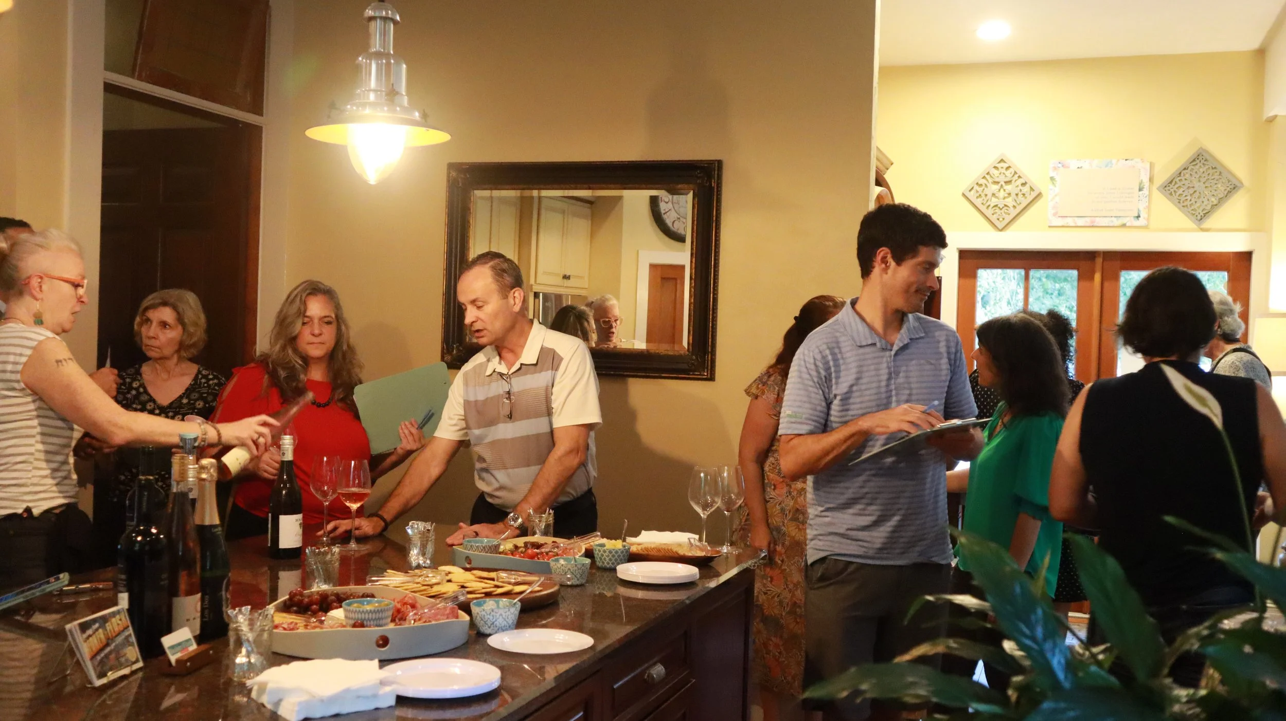 People gathered around a kitchen island with drinks and food, some sitting and some standing, in a well-lit room during a social gathering.