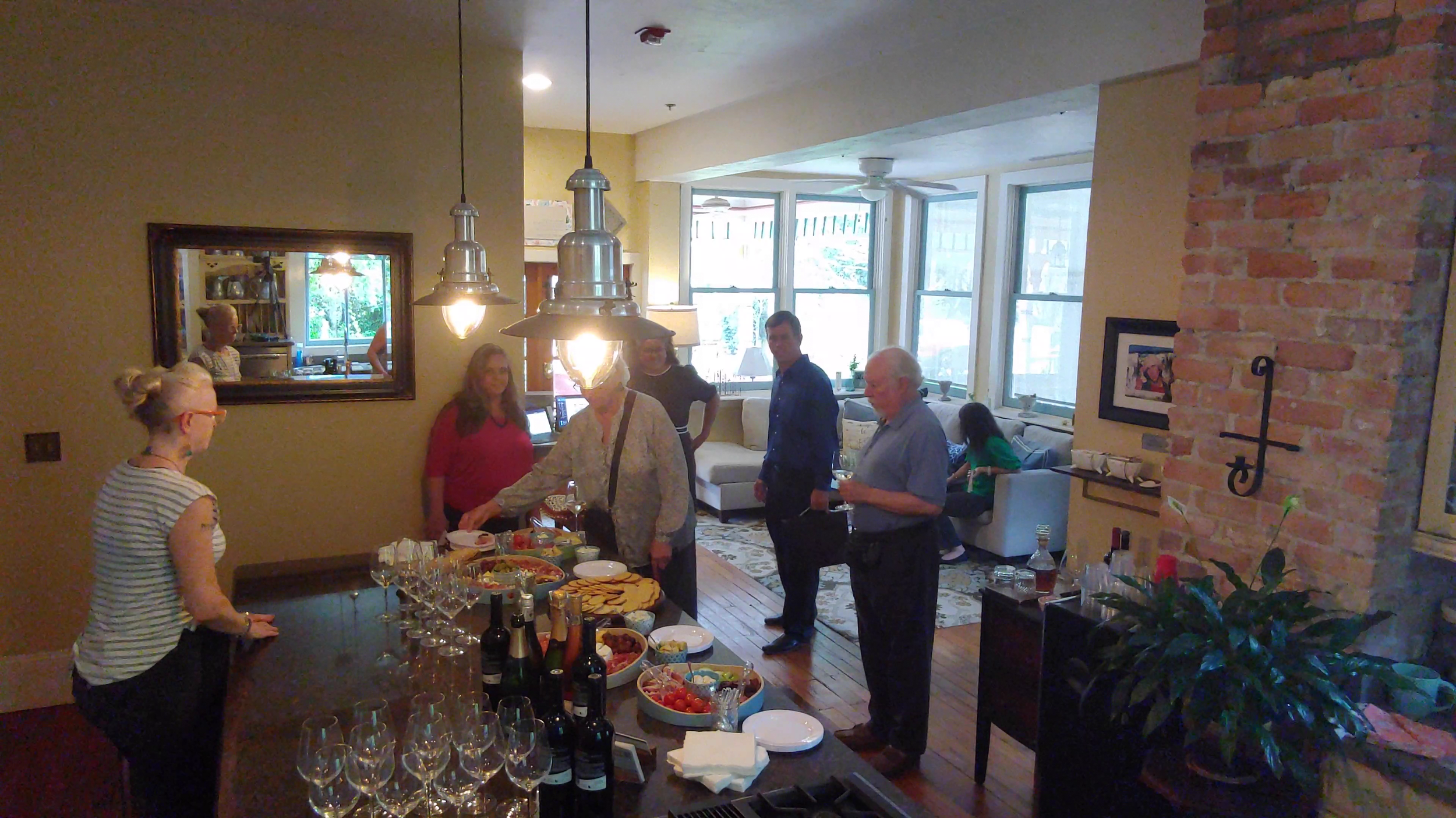 People gathered in a cozy living room and dining area with large windows and a brick wall, enjoying food and drinks at a gathering.