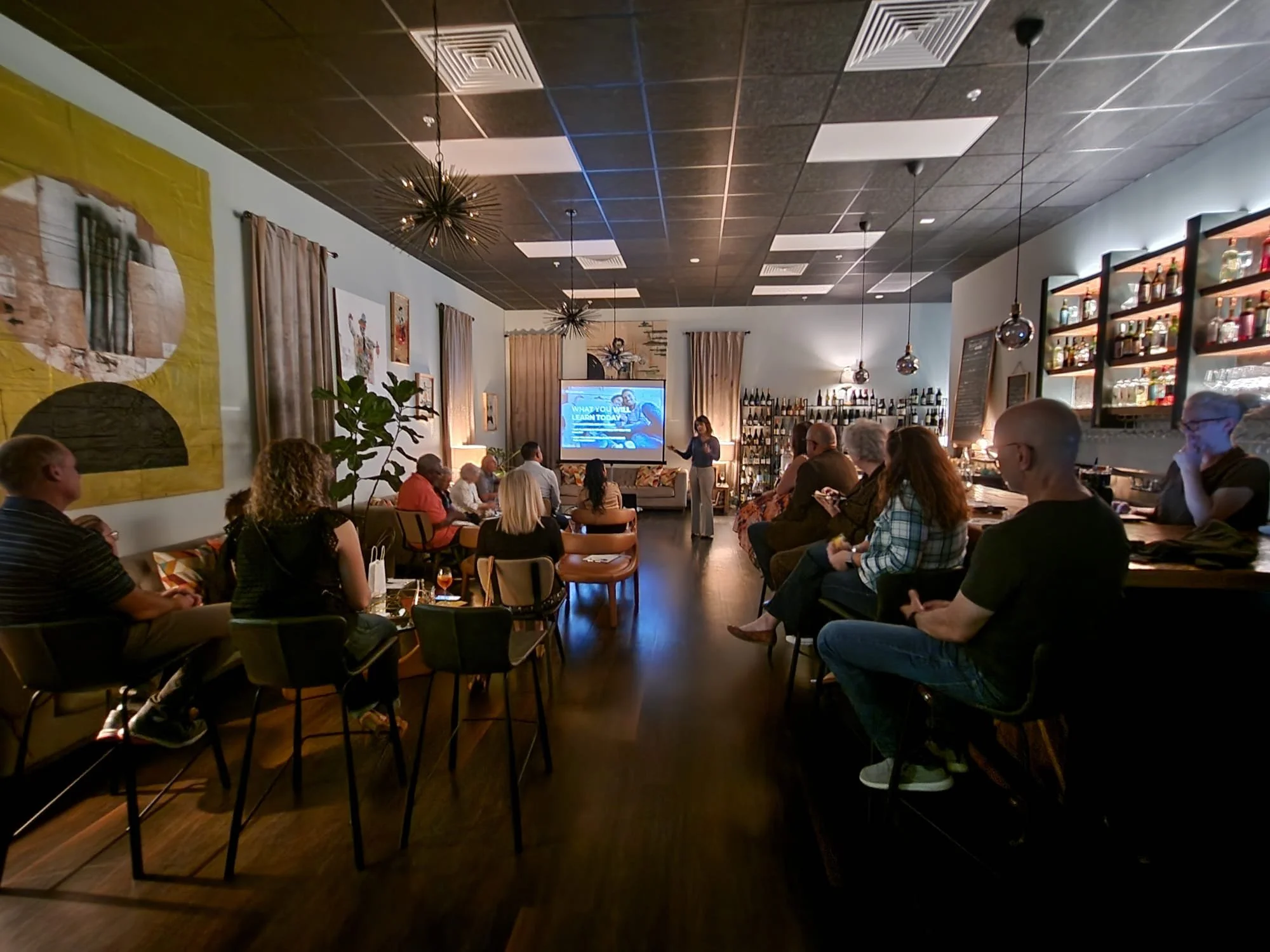A group of people attending a presentation or event in a cozy, modern bar or lounge. The presenter is standing near a screen at the front of the room, with attendees seated and looking towards her.