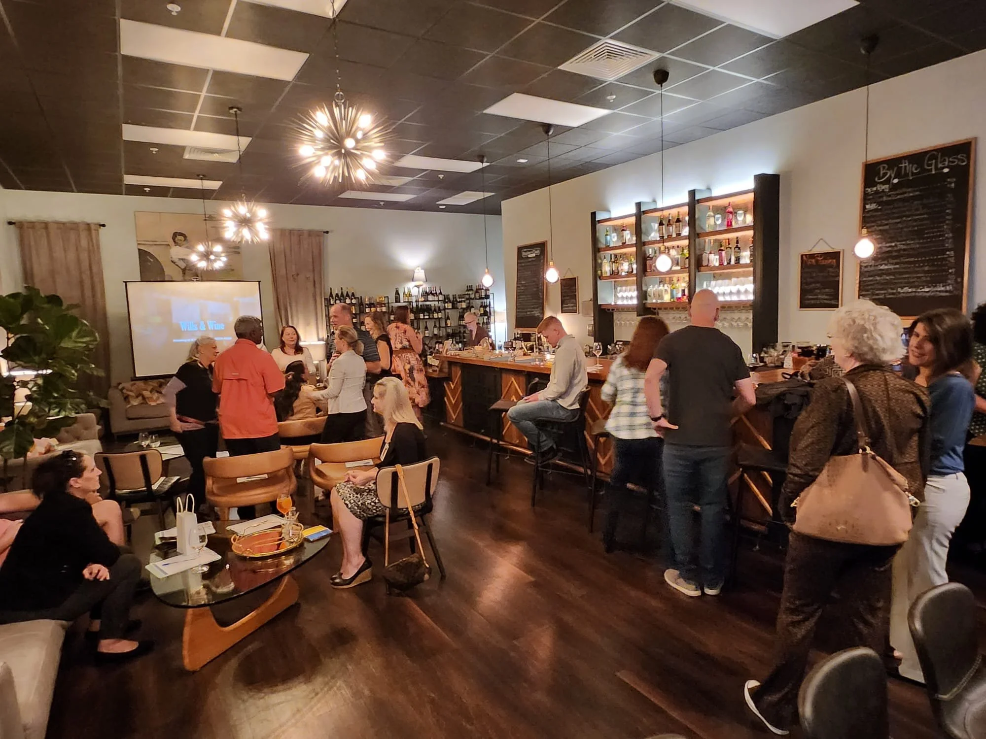 People socializing in a cozy bar or lounge with warm lighting, a large bar counter, shelves filled with bottles, and a television screen showing a presentation or video, in a dimly lit indoor setting.