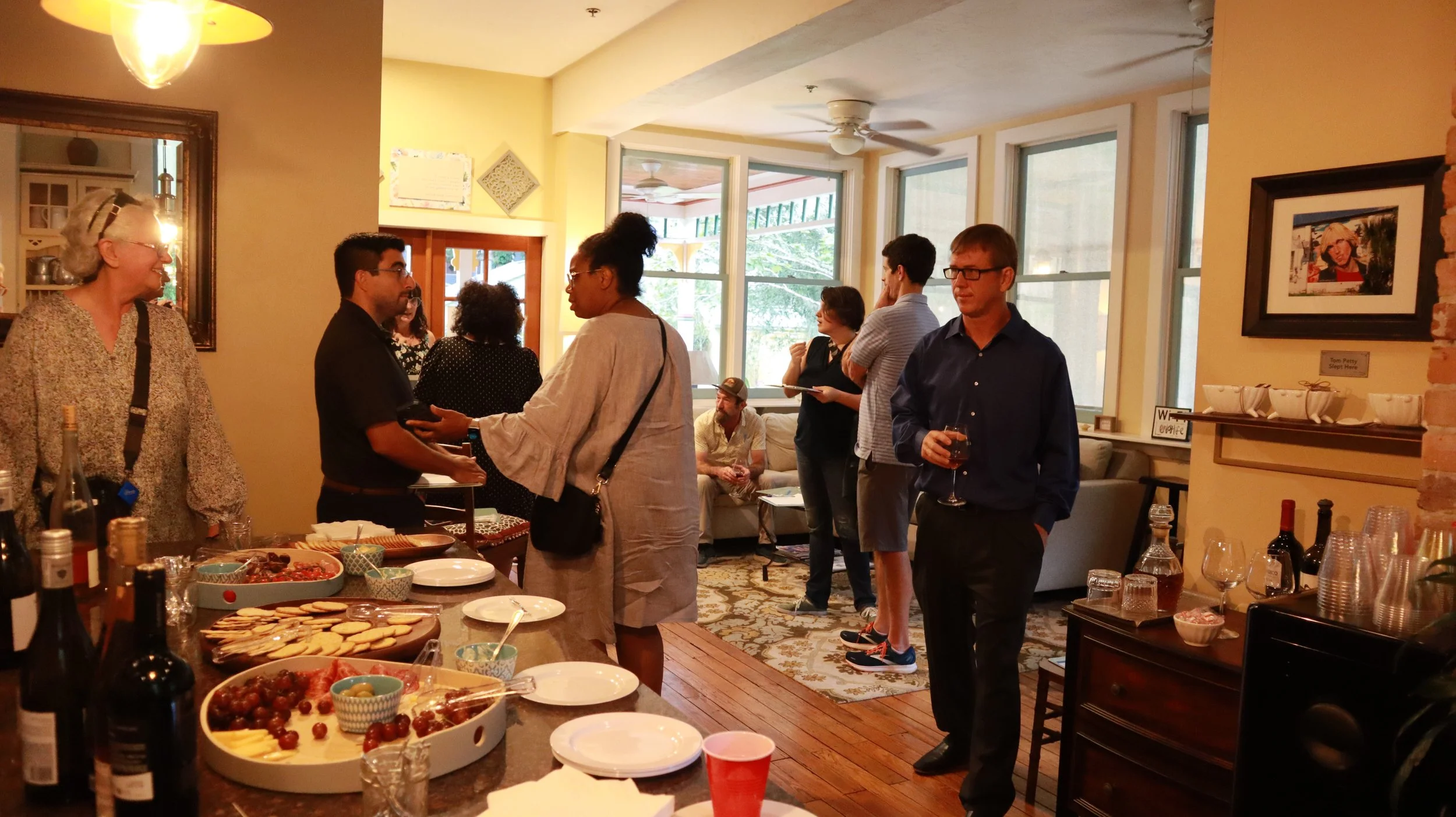 People gathered in a living room during a social event, with a table of snacks and drinks, some guests standing and chatting, and others sitting on a couch, with windows in the background.