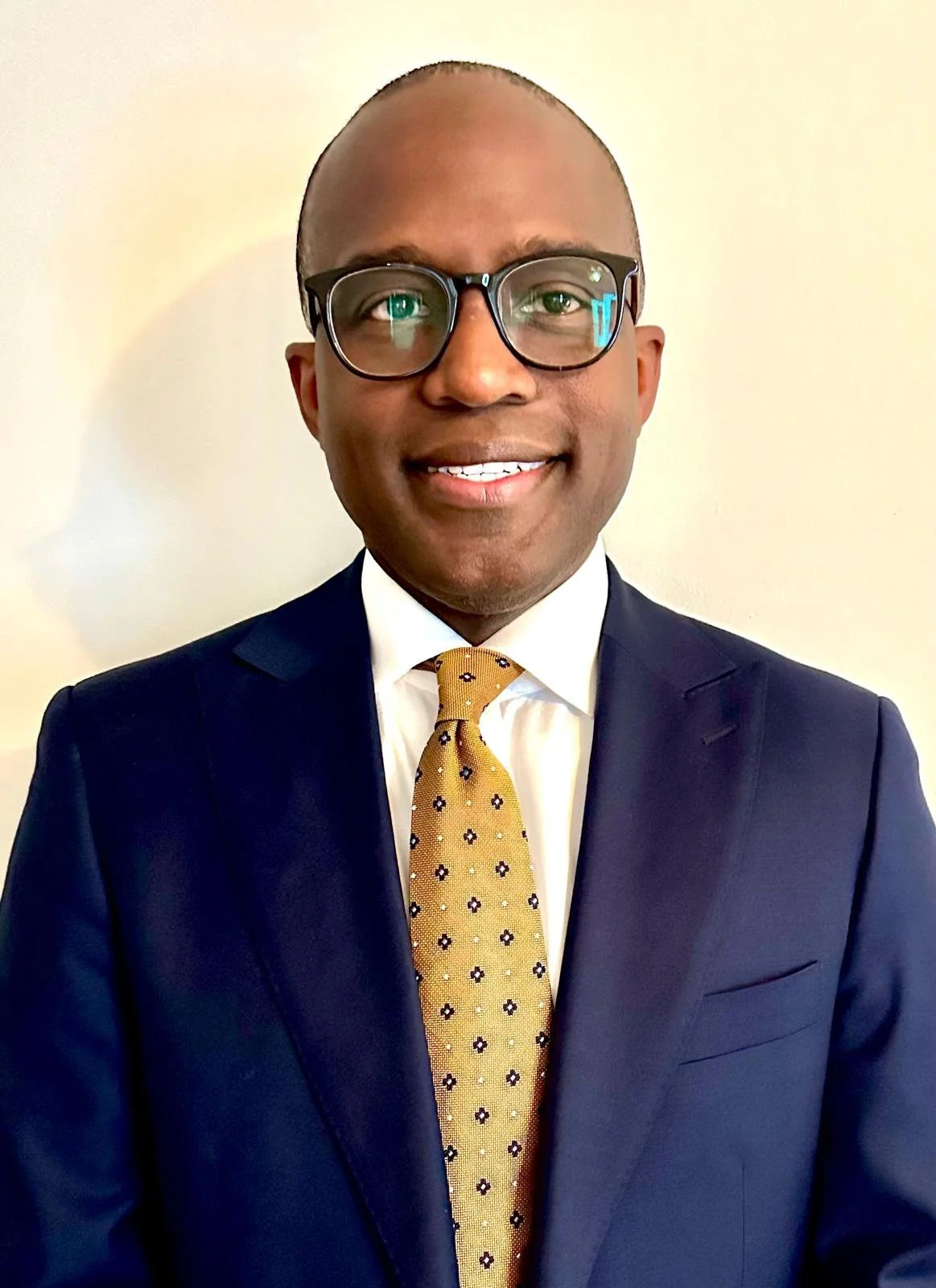 A man in a navy blue suit, white shirt, and yellow patterned tie, wearing glasses, smiling with a neutral background.