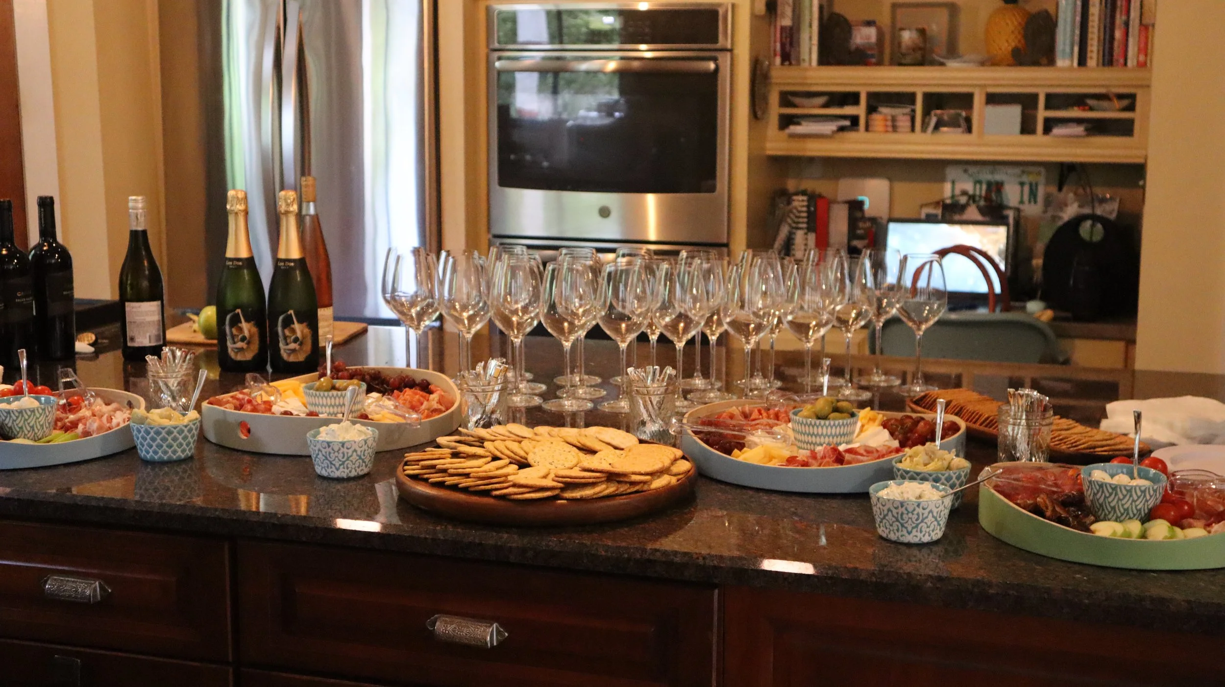 A kitchen island is set up with various appetizers, glasses of wine, and bottles of champagne, with a refrigerator and cabinetry in the background.