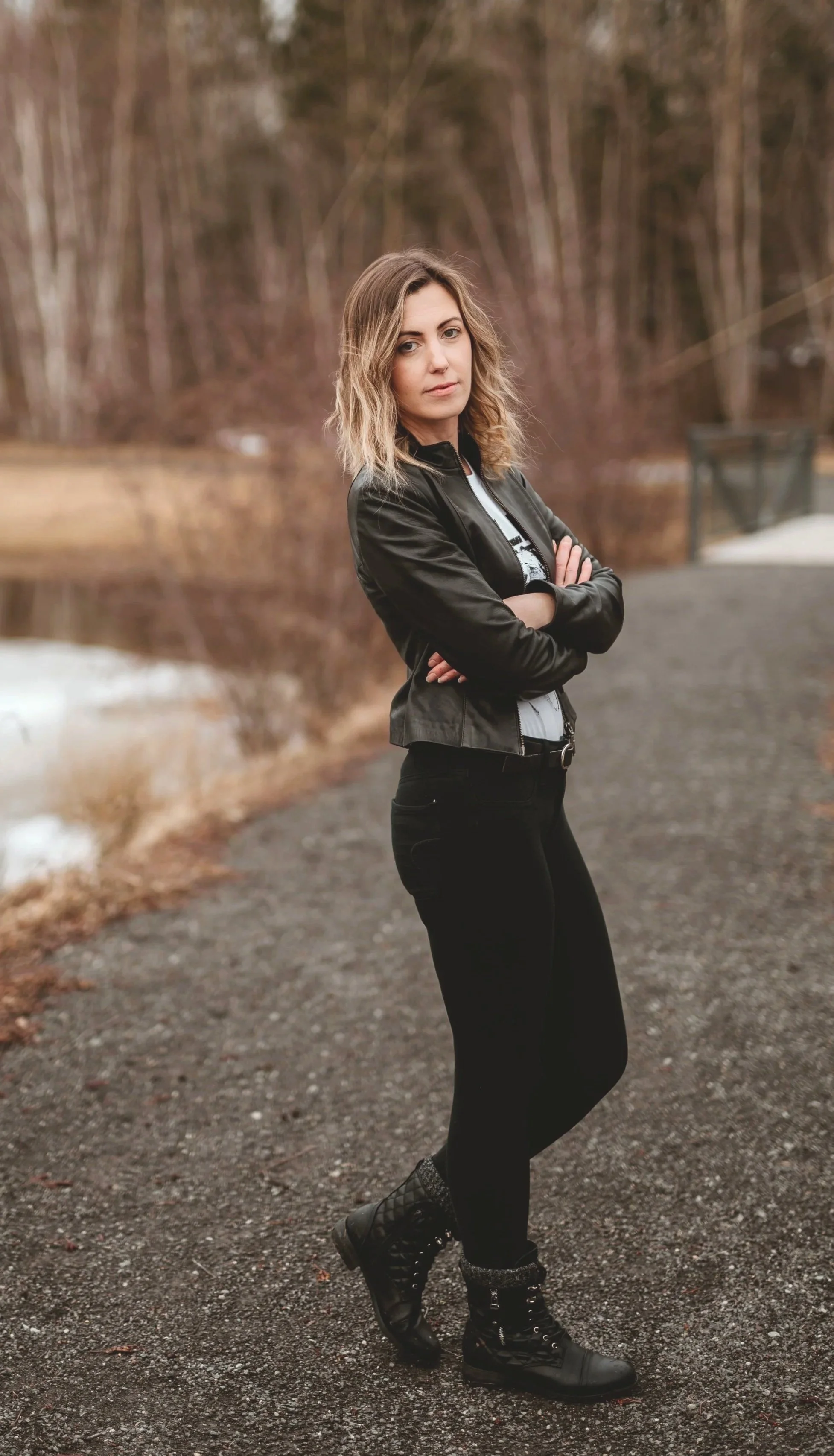 A woman standing on a gravel path near a lake, wearing a black leather jacket, black pants, and black combat boots, with her arms crossed, in an outdoor setting with trees in the background.