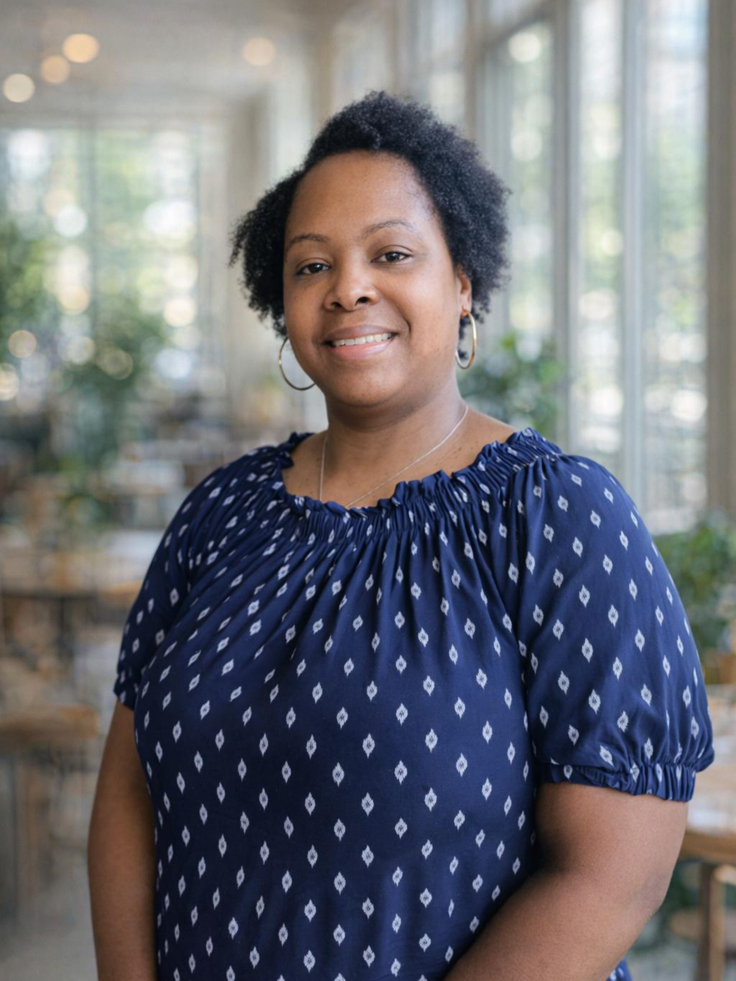 A woman with short curly hair wearing a navy blue blouse with white diamond patterns and hoop earrings, standing in a bright indoor space with large windows and blurred greenery outside.