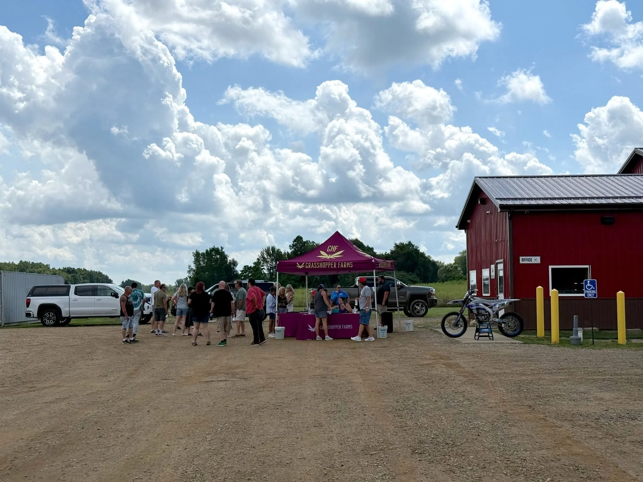 People gathered outdoors near a pink tent and red barn, with a dirt ground and blue sky with large white clouds overhead.