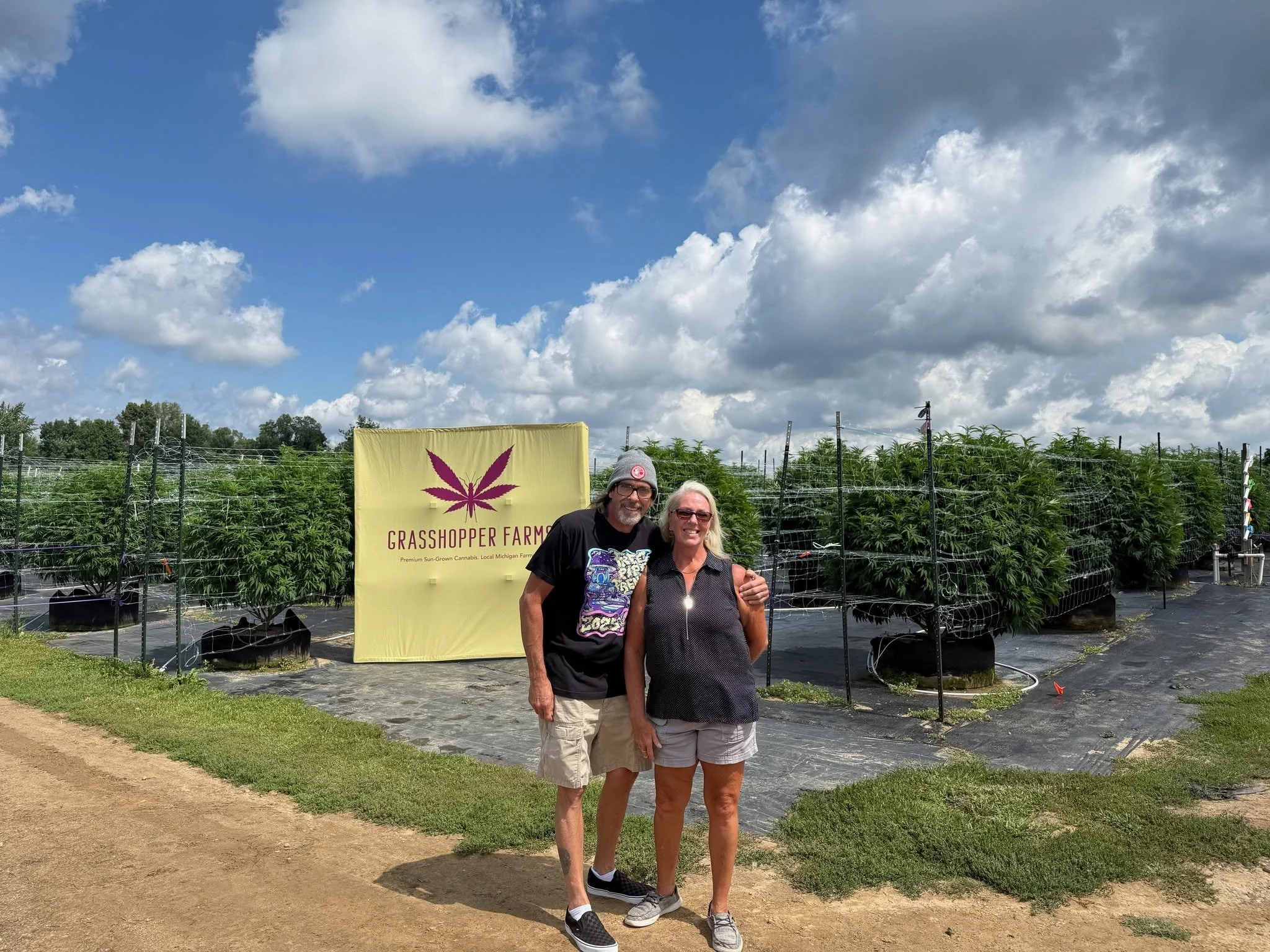 Two people standing outside near a cannabis farm with a sign that reads 'Grasshopper Farms' and depicts a purple cannabis leaf. The sky is partly cloudy, and there are rows of cannabis plants behind them.