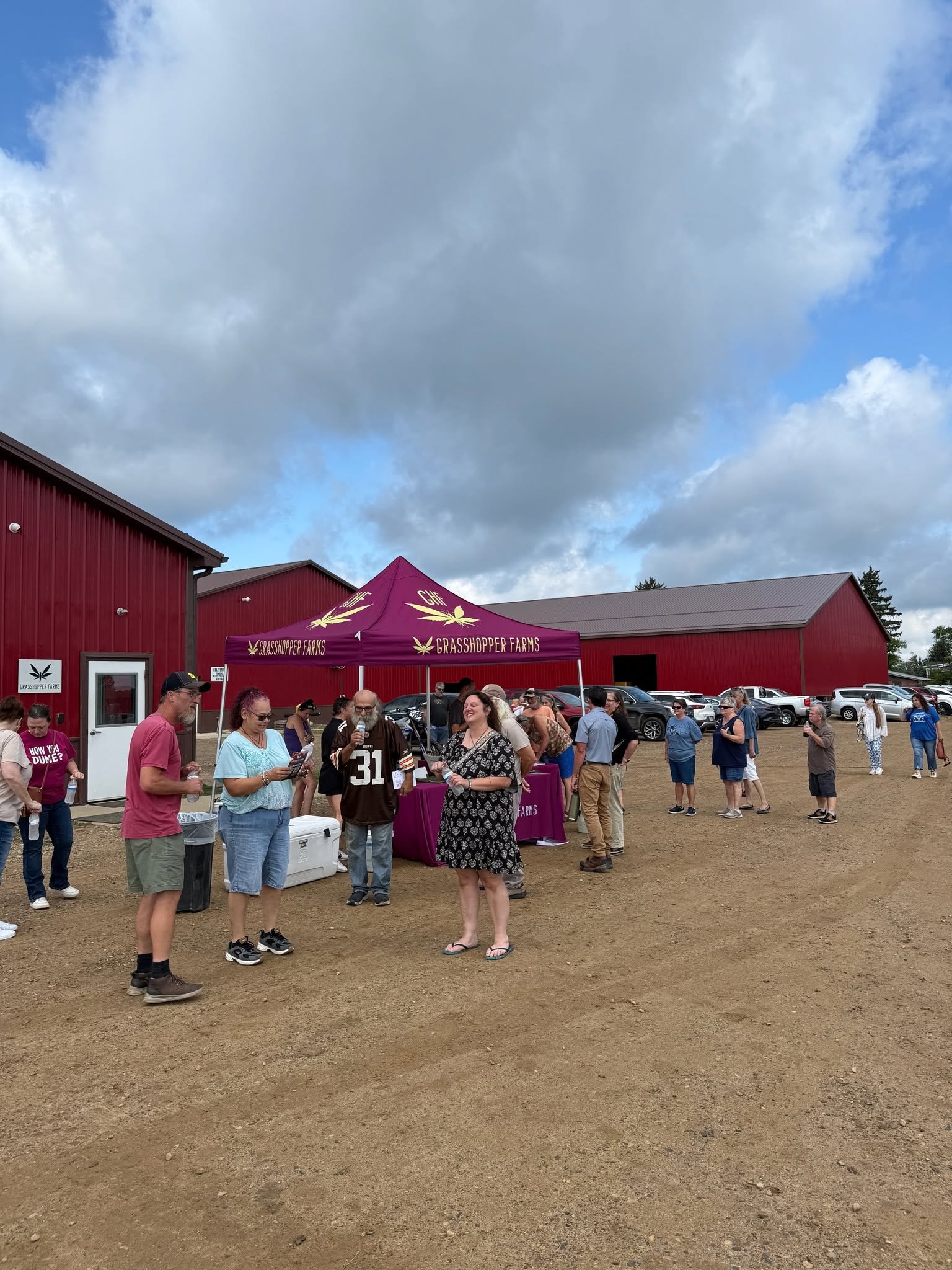 People gathered outdoors near red buildings under a cloudy sky, with a purple canopy labeled Grasshopper Farms.