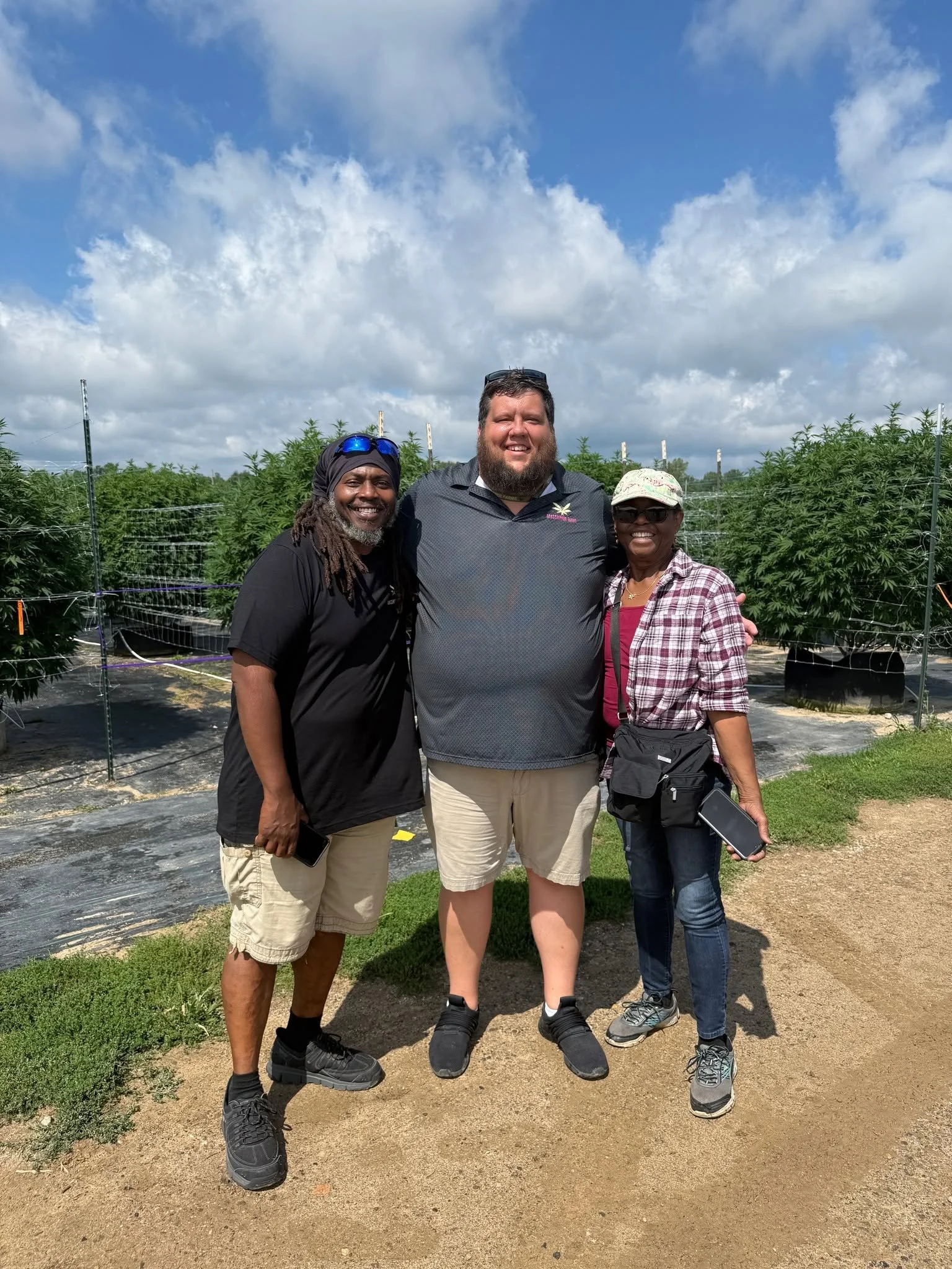 Three people standing outdoors in a garden, smiling, with plants and a partly cloudy sky in the background.