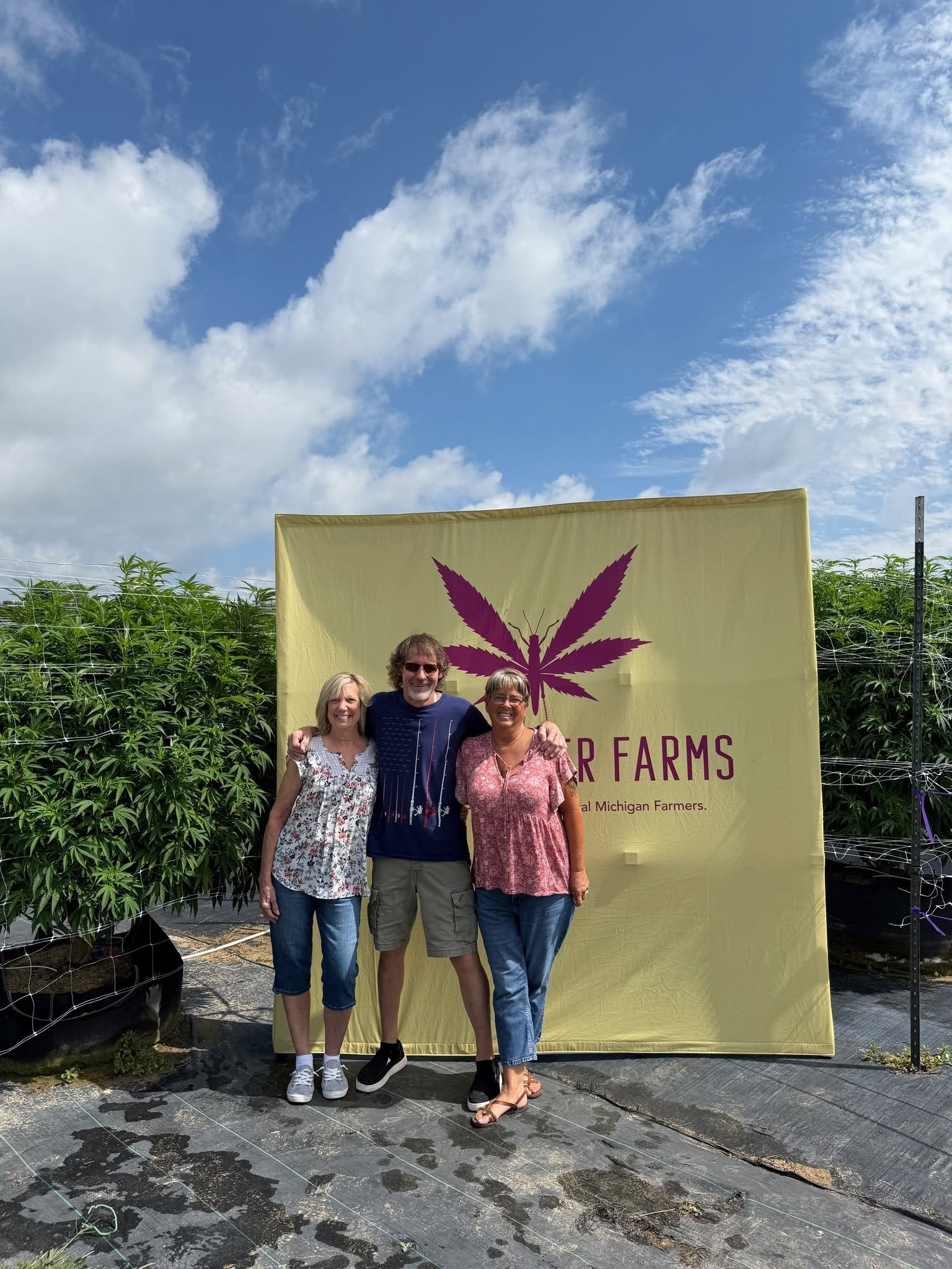 Three people standing in front of a yellow banner with a purple cannabis leaf graphic and the words 'Far Farms' at a cannabis farm, with green plants behind them and a partly cloudy blue sky above.