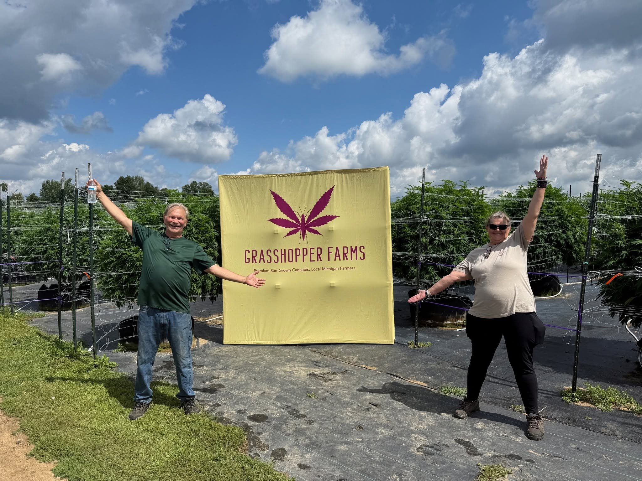 Two smiling people standing outdoors on a cloudy day in front of a sign that reads "Grasshopper Farms" with a purple marijuana leaf logo, surrounded by rows of cannabis plants.