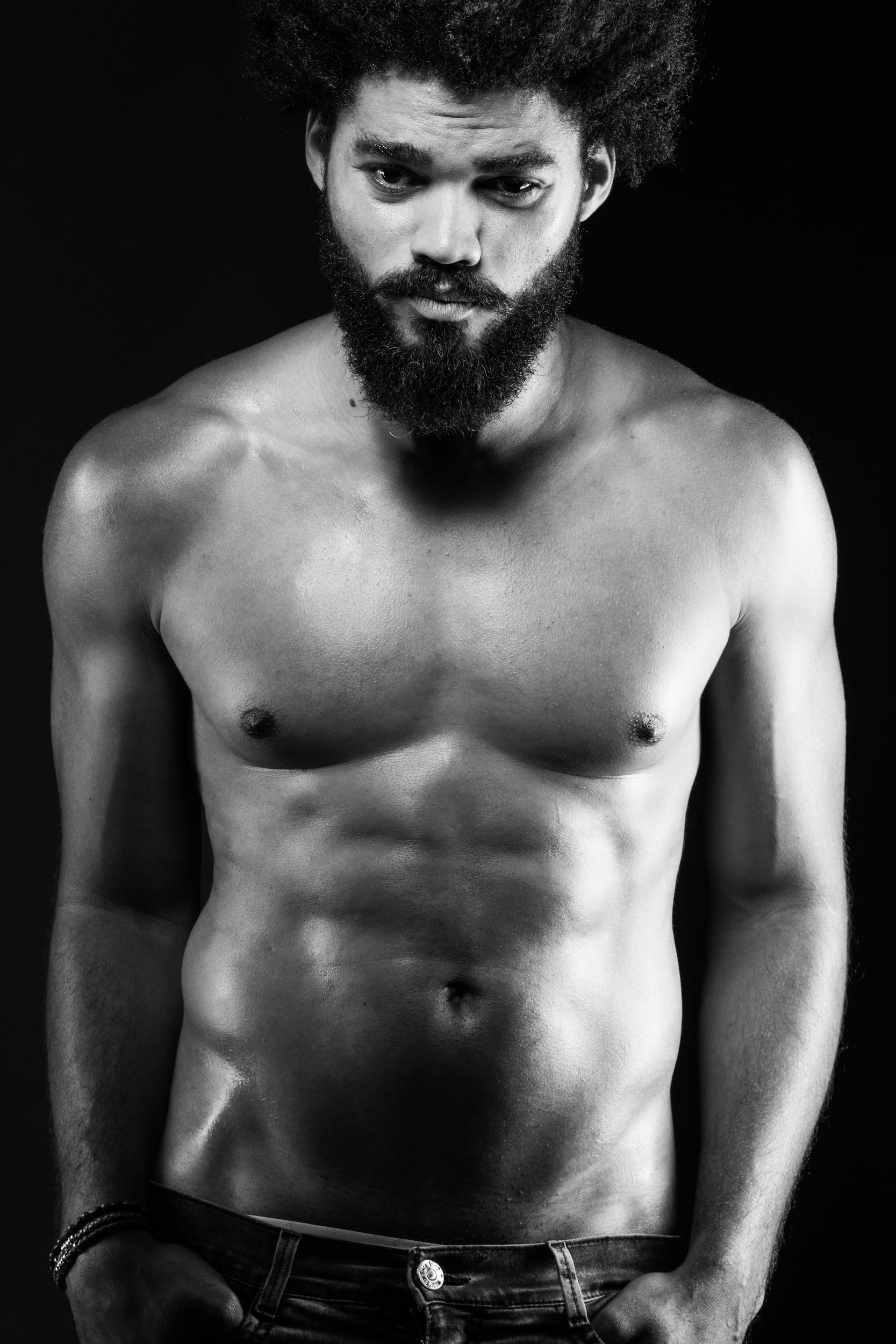 Black and white portrait of a shirtless man with a beard and curly hair, looking directly at the camera, against a dark background. (Sex, intimacy, romance, aphrodisiac, tantra, consuming, immersive, horny, couples, relationship, relational)