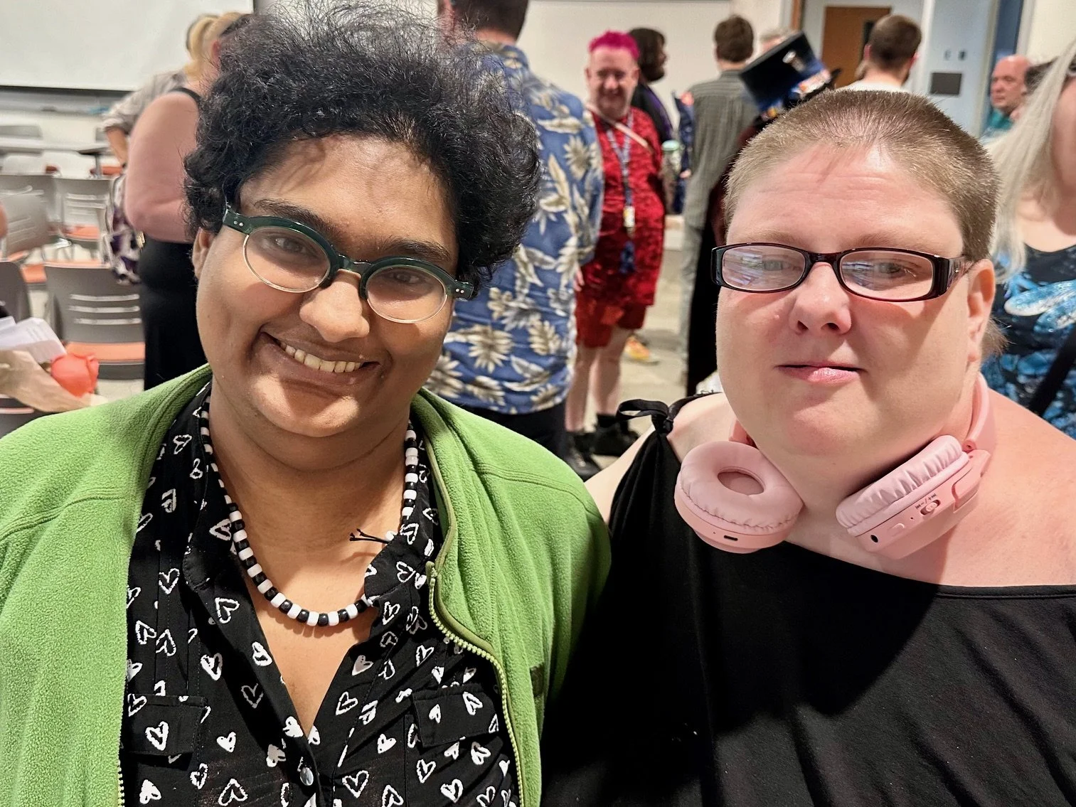 Chandra wears a lime green jacket, glasses, and a smile, and stands next to SuzyQ who is pictured also wearing glasses, a black top and pink headphones around her neck. They stand in Purce Hall on the campus of The Evergreen State College