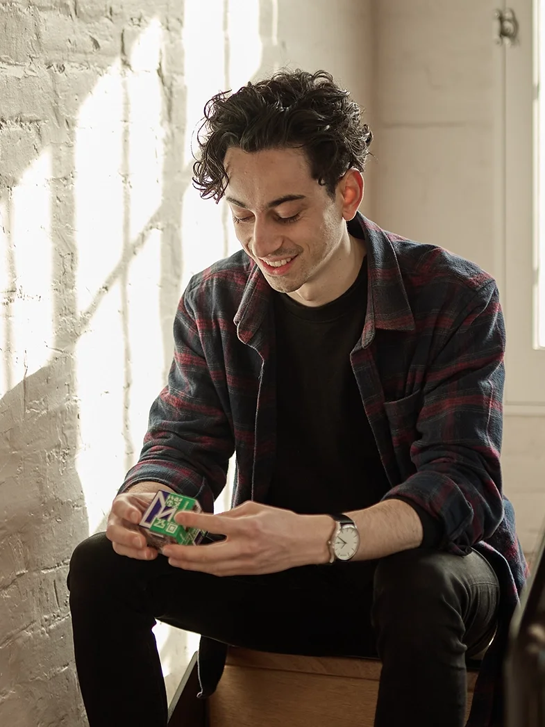 A young man with dark, curly hair, wearing a black t-shirt and a plaid flannel shirt, sitting on a wooden surface, smiling while holding a deck of UNO cards in his hands, near a brick wall with sunlight coming through a window.