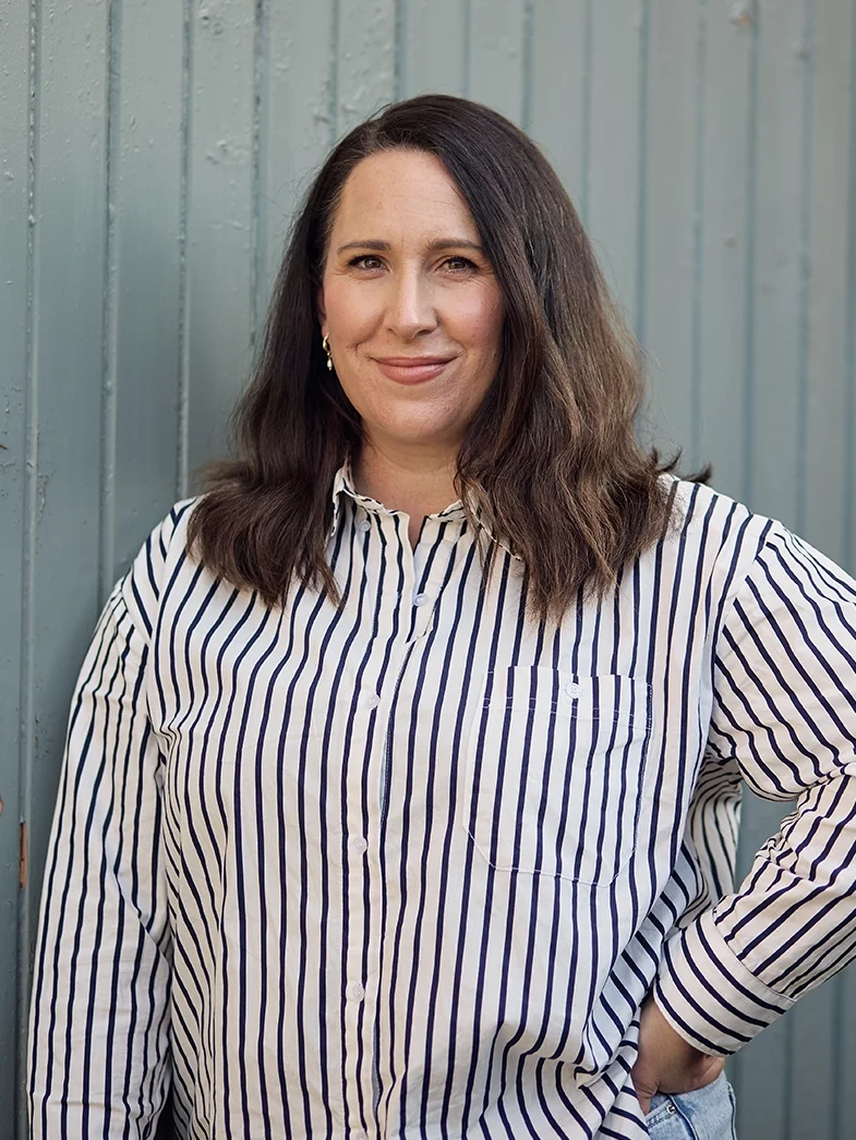 A woman with shoulder-length dark hair, dressed in a white shirt with black vertical stripes, standing outdoors in front of a blue wooden fence, smiling slightly with her right hand on her hip.