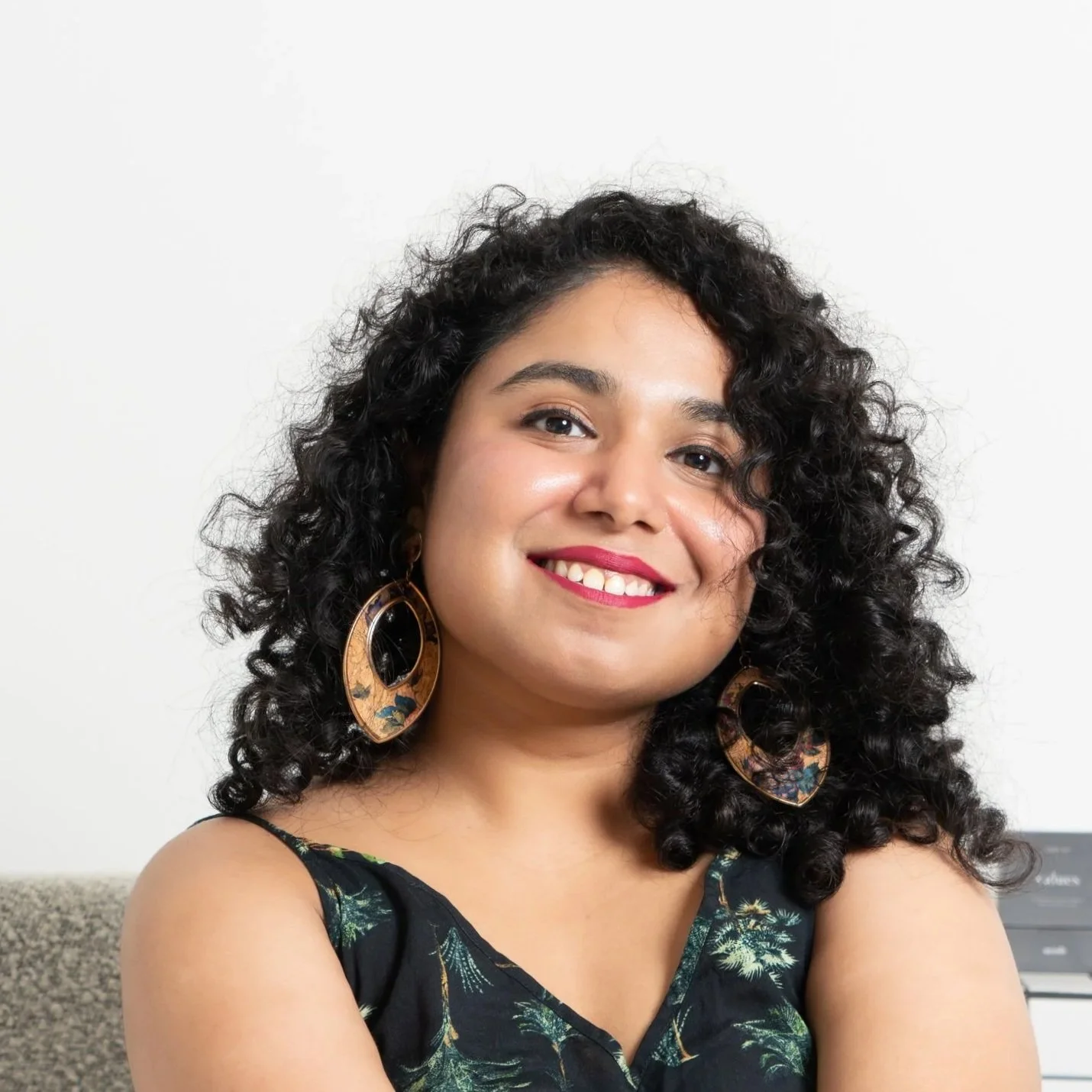 A woman with curly black hair, wearing a black top with a floral pattern, large floral earrings, and bright red lipstick, smiling in front of a plain white background.