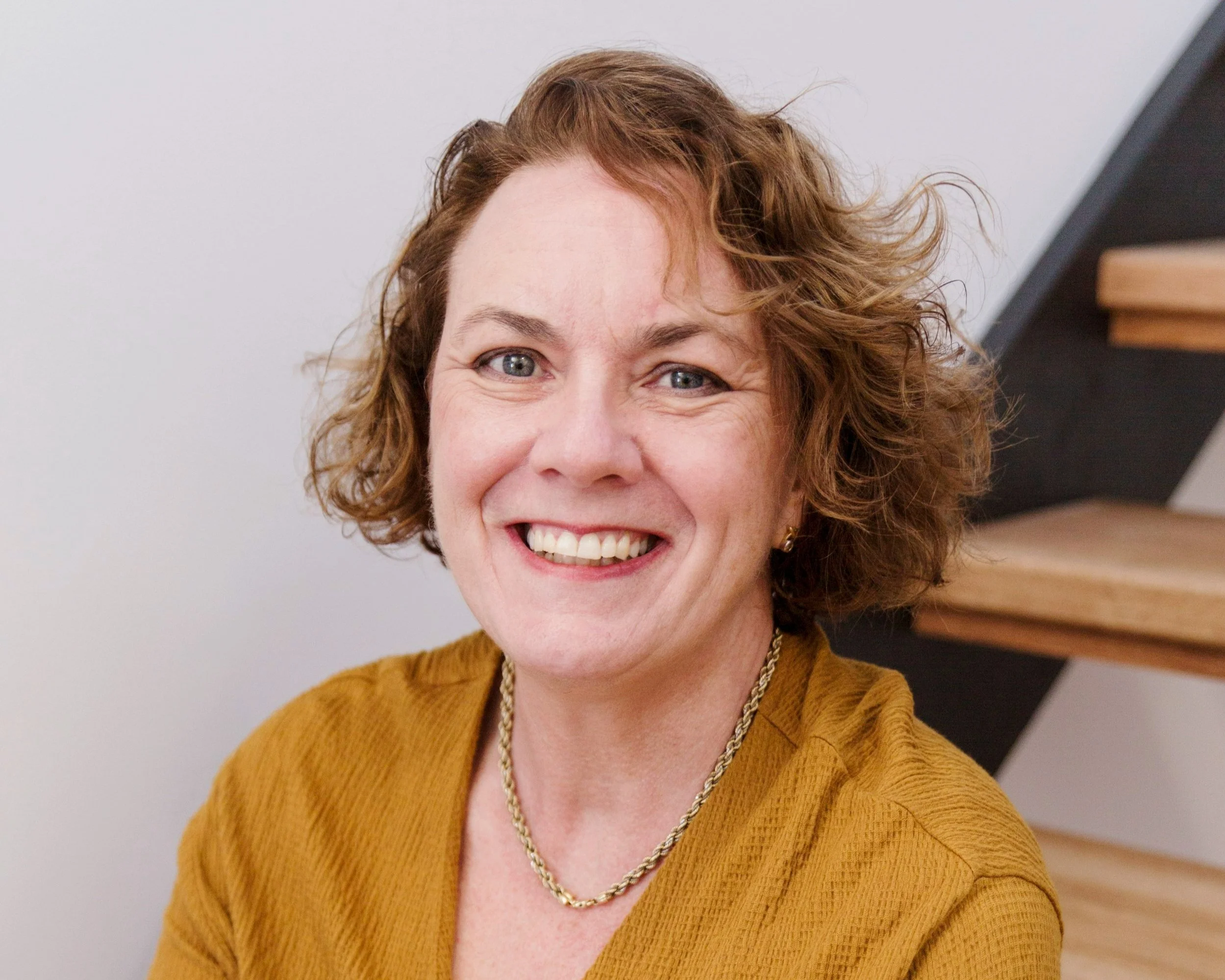 A woman with curly, shoulder-length hair and blue eyes, smiling at the camera, wearing a mustard-colored top and gold jewelry, sitting on wooden stairs with a white wall in the background.