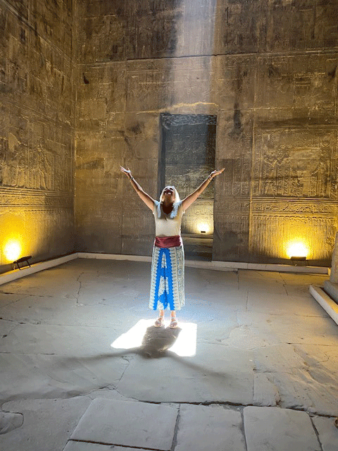 Woman standing in a shaft of light in an Egyptian temple with arms outstretched and looking toward the sunlight.