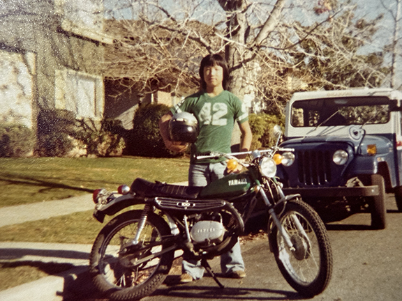Young Korean man in a green t-shirt with the number 42 on the front. He holds a helmet under his right arm and stands behind a green Yamaha motorcycle in a residential neighborhood.