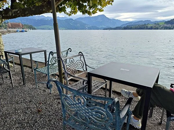 Cafe tables on a gravel seating area right against the lake with mountains in the distance.