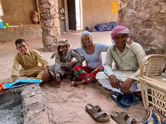 A blonde woman sitting on the sandy ground with a man and two boys. One of the boys and the man wear a head wrap. The structures around them are made of sandy colored stone.
