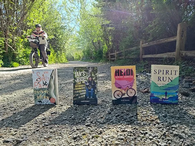 A trail with a woman riding by on a bicycle. It's sunny and the trees along the trail are green. On the path are four books: The Salt Path by Raynor Winn, Ian's Ride by Karen Polinsky, Heidi Across America by Heidi Beierle and Spirit Run Noe Alvarez