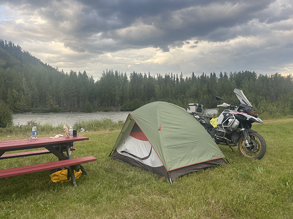 Mountain setting by water. Evergreens rim the water, the sky is cloudy buy bright. On the grass in the foreground is a picnic table with some water bottles, a tent set up, and a motorcycle parked next to the tent.
