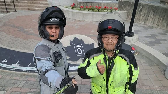 Two men in motorcycle gear: Michael in gray on the left and Karl in safety yellow on the right making a thumb's up. Between them is a 0km marker in a bricked plaza.