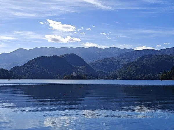 Blue sky, blue lake, completely forested hills and mountains in the distance. A church peeks out of the trees on a small island at the far end of the lake.