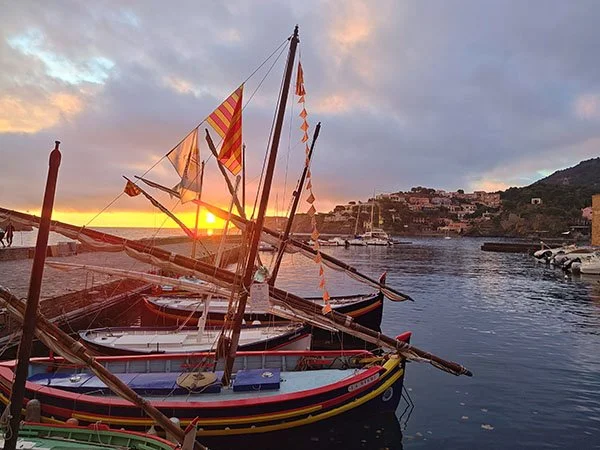 Colorful boats with short masts and stowed sails moored in a bay. A yellow stripe of sunrise lightens the sky with pink, purple, and blue clouds above.