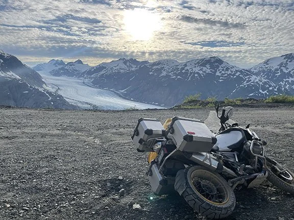 Motorcycle laying on the gravel. In the distance is a glacier between two mountain ranges and a radiant sun behind light clouds.