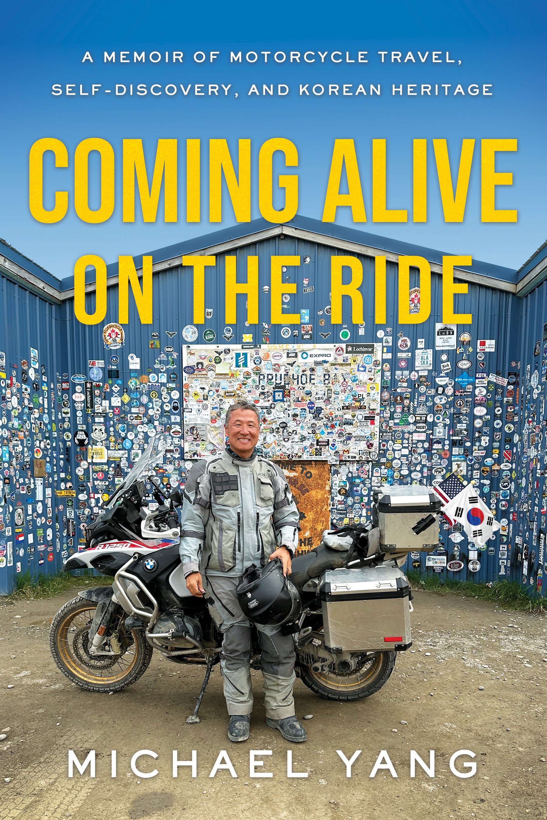 Korean man standing in front of his parked motorcycle in front of a building that says Prudhoe Bay. The building is covered with stickers. The book's subtitle is A memoir of motorcycle travel, self-discovery, and Korean heritage.