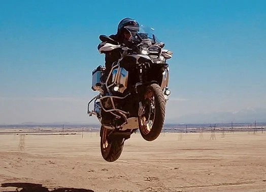 Motorcycle in air with a determined rider. The land as far as can be seen is dry dirt dotted with electrical transformers. Faint mountains at the horizon. Bright blue sky.