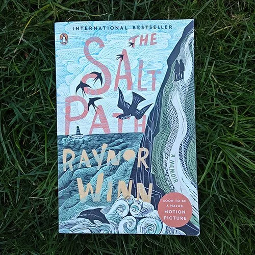 The Salt Path by Raynor Winn. The cover is a block print style illustration with a couple on a coastal path, waves at the bottom with a dolphin, and swifts and a peregrine falcon in the sky. International Bestseller.