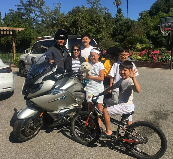 Michael Yang in his motorcycle gear with his motorcycle. A short woman is next to him and his four children: three boys and a girl. The youngest boy in the foreground on a bicycle waves to the camera, and the girl holds a small white dog.