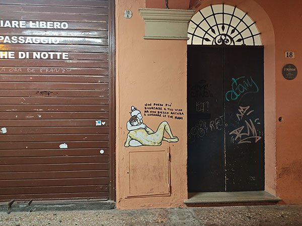 Light orange building. A black door with decorative iron in the arch above the black. 18 To the left of the door is a closed rolling garage door. Between the two is light orange wall with a painting of a naked man and woman embracing.