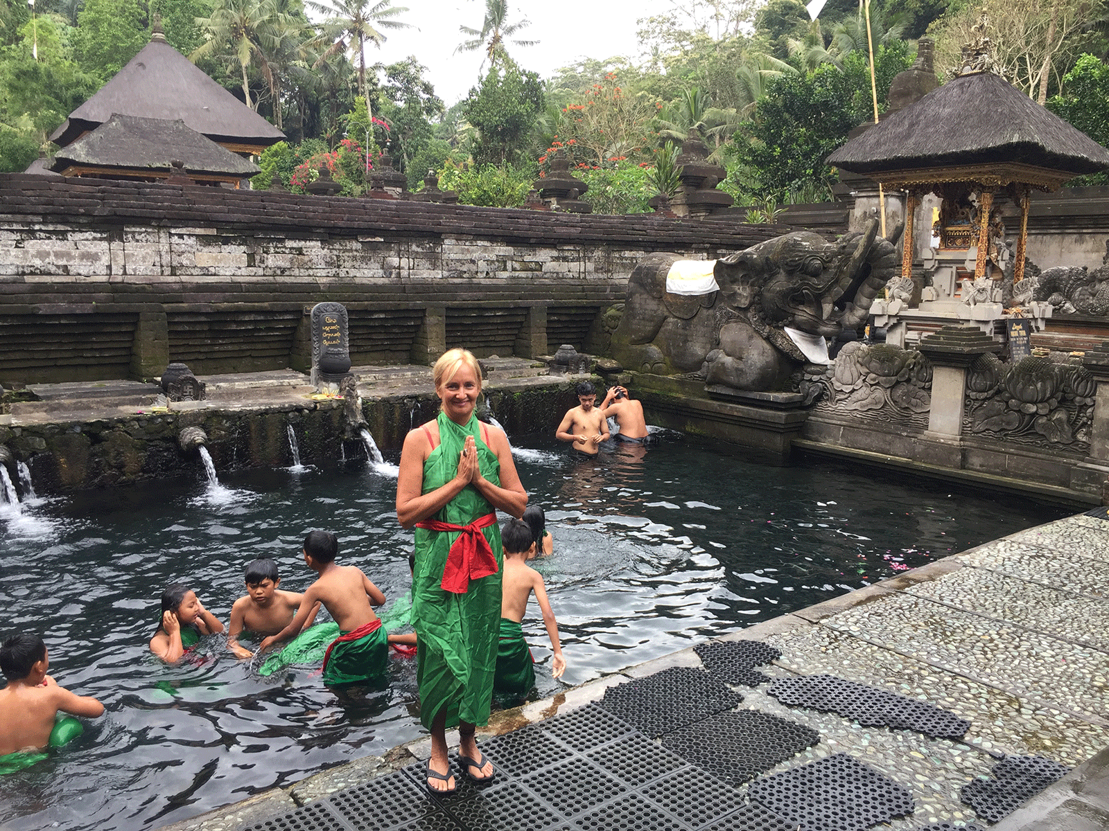 Blonde woman with hands in prayer position wearing a long green shift with a red tie around the waist. She's at a pool where several children are in the water.