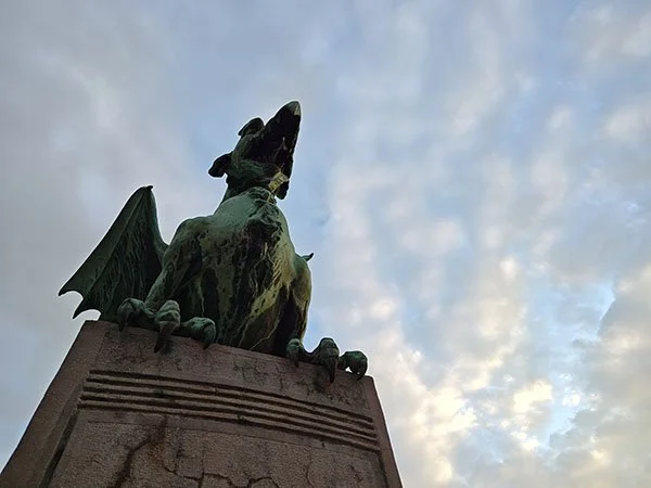 A view looking up into a green patina dragon sculpture. The dragon's mouth is open, it's wings are visible, and it's claws on both front feet are curled over the edge of the pedestal upon which it sits.