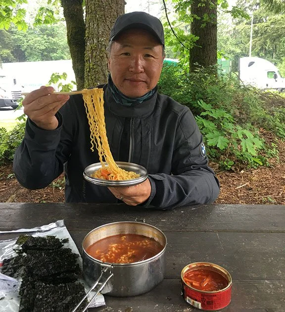 A Korean man in a ball cap and motorcycle jacket at a rest area picnic table smiling and holding a wad of ramen noodles with chopsticks. On the table is a camp pot of red soup and a package of nori snacks.