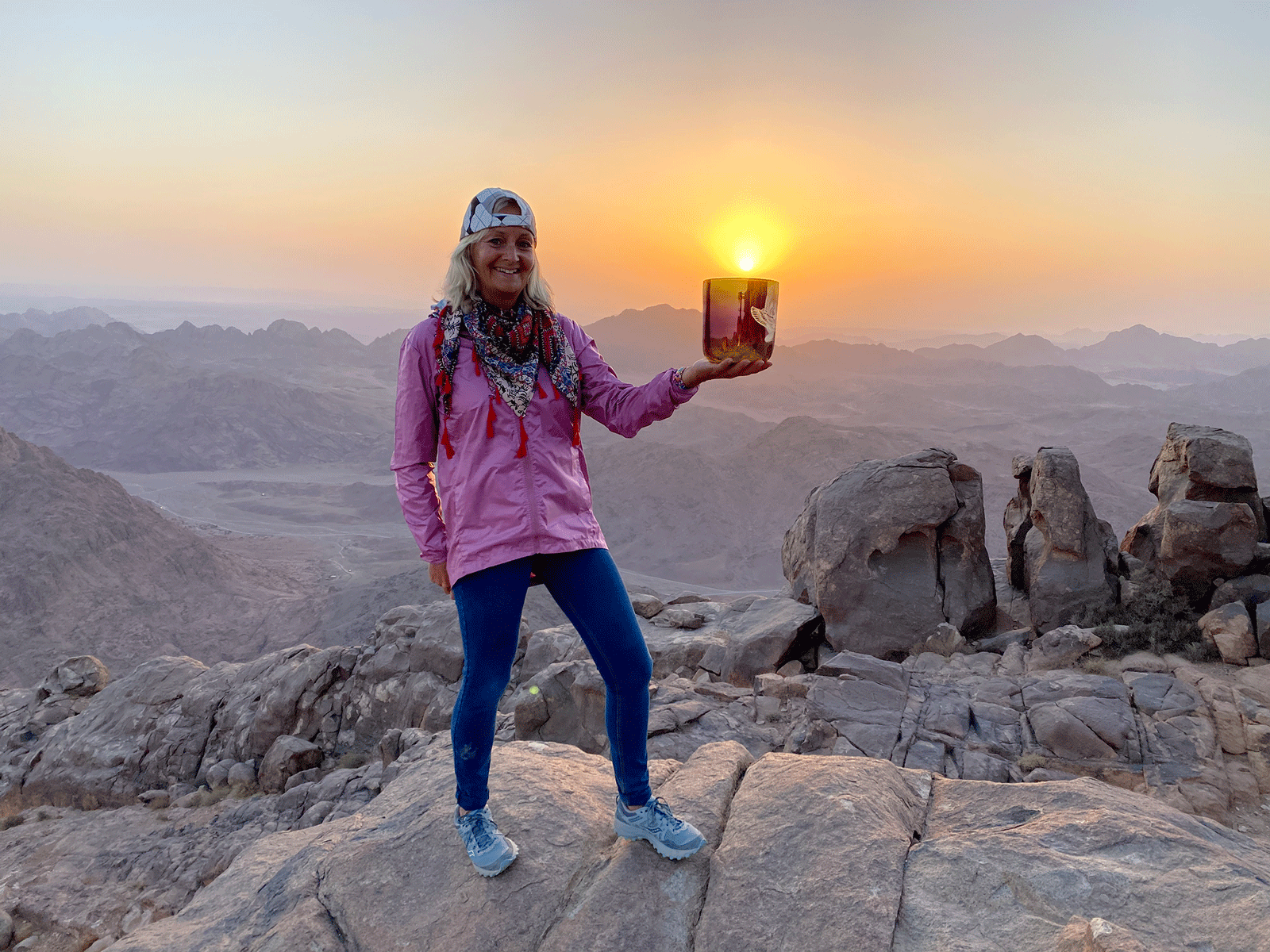A woman standing atop a desolate, rocky place holding a reflective vessel from which the sun is rising.