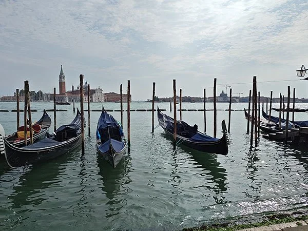 Four gondolas tied to posts in the Grand Canal. A spire with other buildings is visible in the distance. The water squiggles with reflections from the boats.