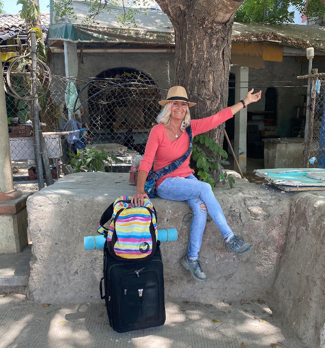 Blonde woman sitting on a wall with a suitcase, rainbow backpack, and yoga mat. She's wearing a fedora and one arm is outstretched, her legs are crossed. Chainlink fence and a tree behind her.
