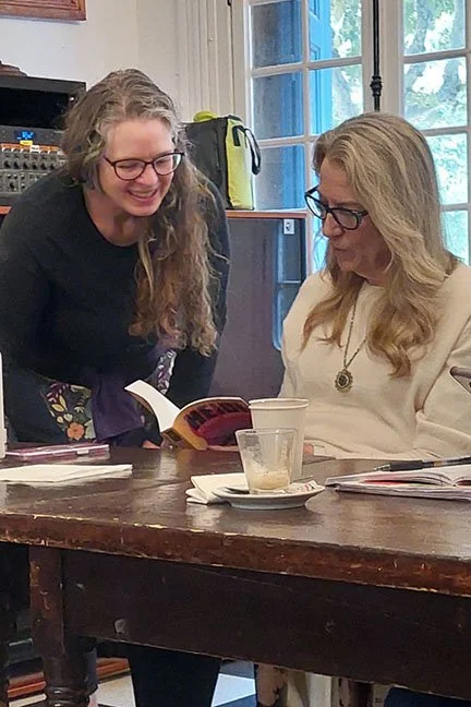 Woman with long curly brown hair smiling next to a woman with blonde hair sitting at a table looking at Heidi Across America book.
