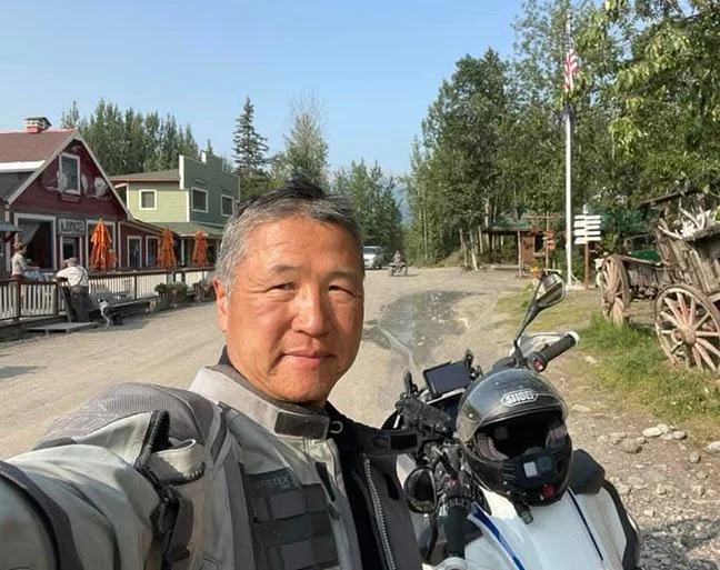 Korean man in a motorcycle jacket. His helmet is resting on a motorcycle just behind him. In the background is a dirt road, rustic town, American flag, wooden wagon, trees, and blue sky.