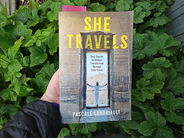 Photo of She Travels book over greenery. The cover depicts a woman in white standing in an Egyptian doorway looking out over water with a mountain behind. The subtitle: real stories of women transformed through solo travel.