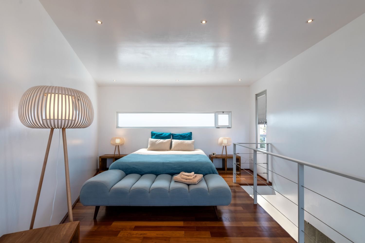 Modernes Schlafzimmer mit minimalistischem Design, Holzboden, großem Bett mit blauer Bettwäsche und Kissen, Fenster, Stehlampe und Nachttischen.